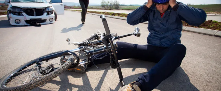 Bicyclists injured in a car accident lying down on the road and holding his head with helmet. A person who is driving car running to helps to injured man.